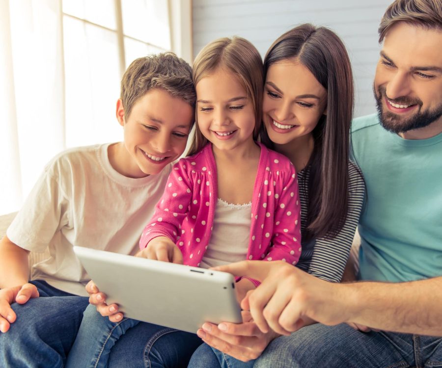 Beautiful young parents, their cute little daughter and son are using a tablet and smiling, sitting on sofa at home